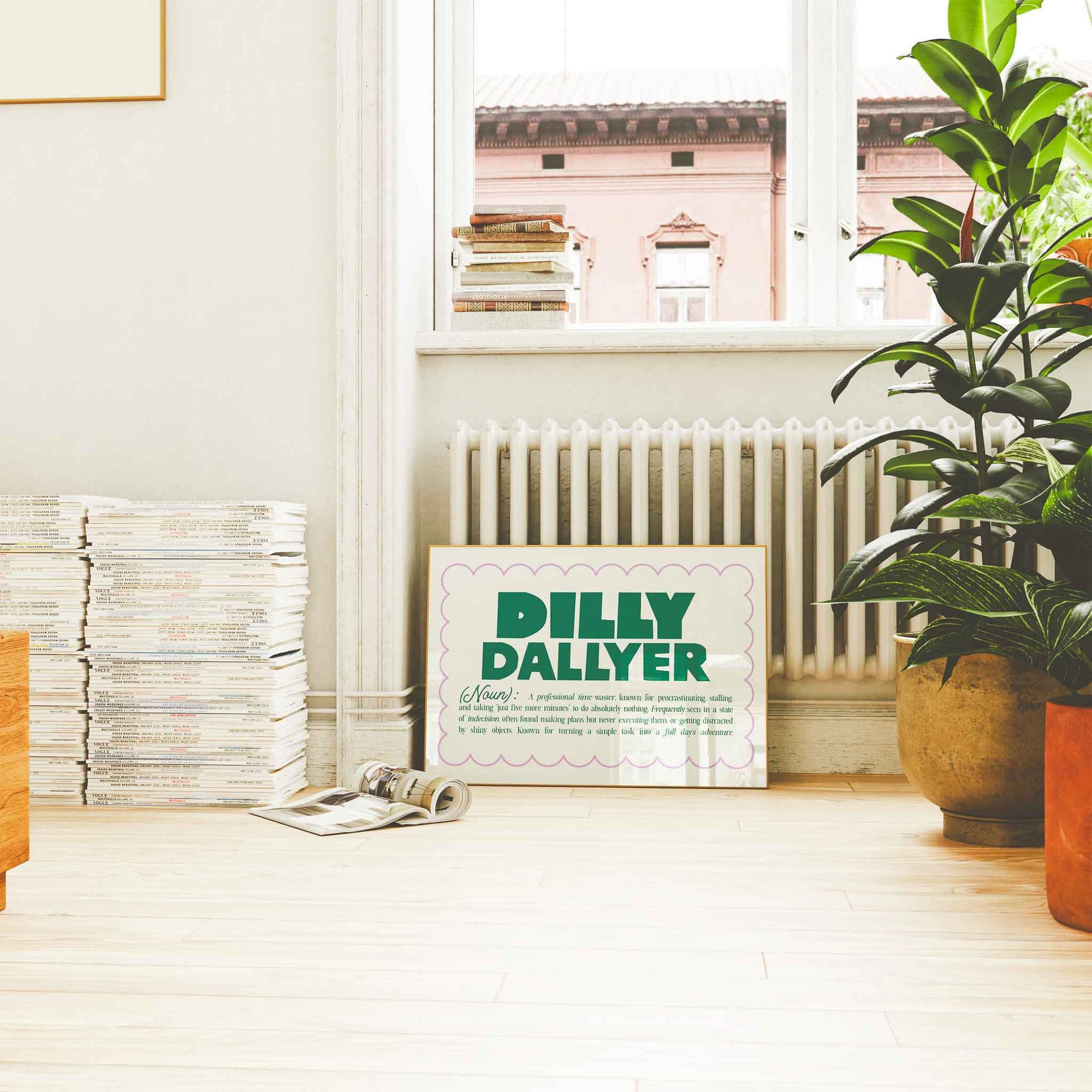 Playful 'Dilly Dallyer' definition print leaning against wall in cozy reading nook with potted plants and stacked magazines.