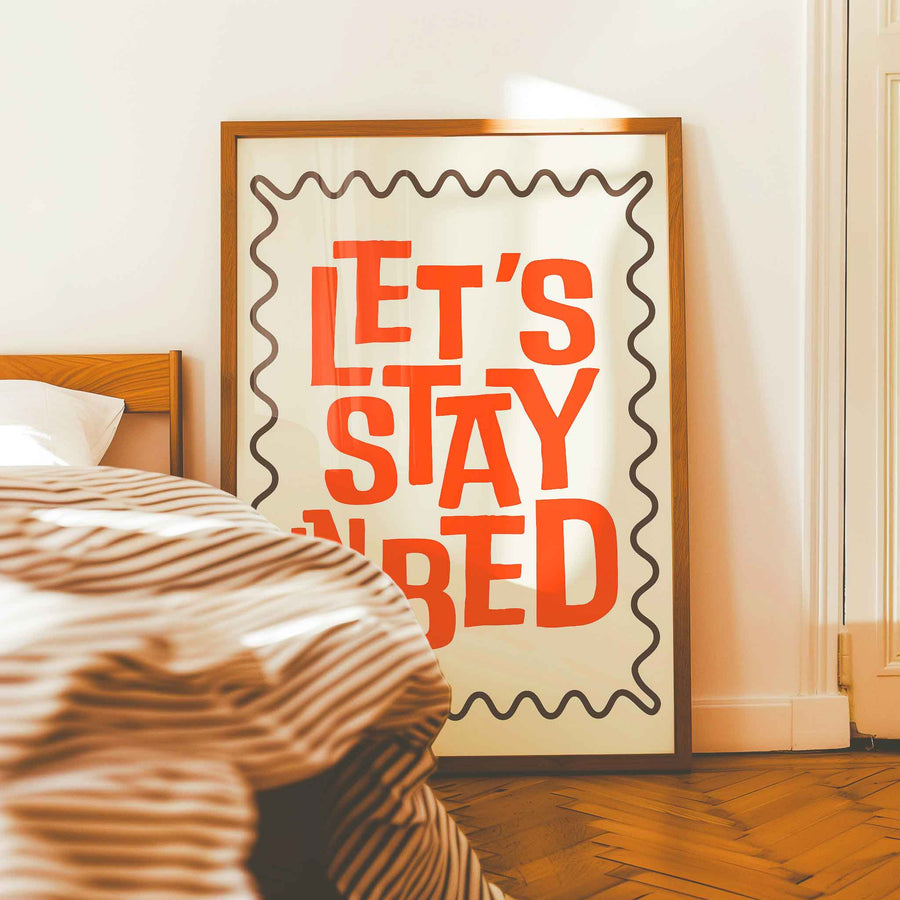 Cozy bedroom wall art print with bold red text that says “Let’s Stay in Bed,” framed and leaning against the wall beside a bed with striped bedding.