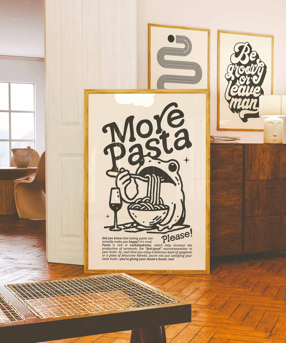 Vintage-style black and white wall art print with a frog eating pasta and a wine glass, framed in a warm-toned interior. Typography says “More Pasta Please.”