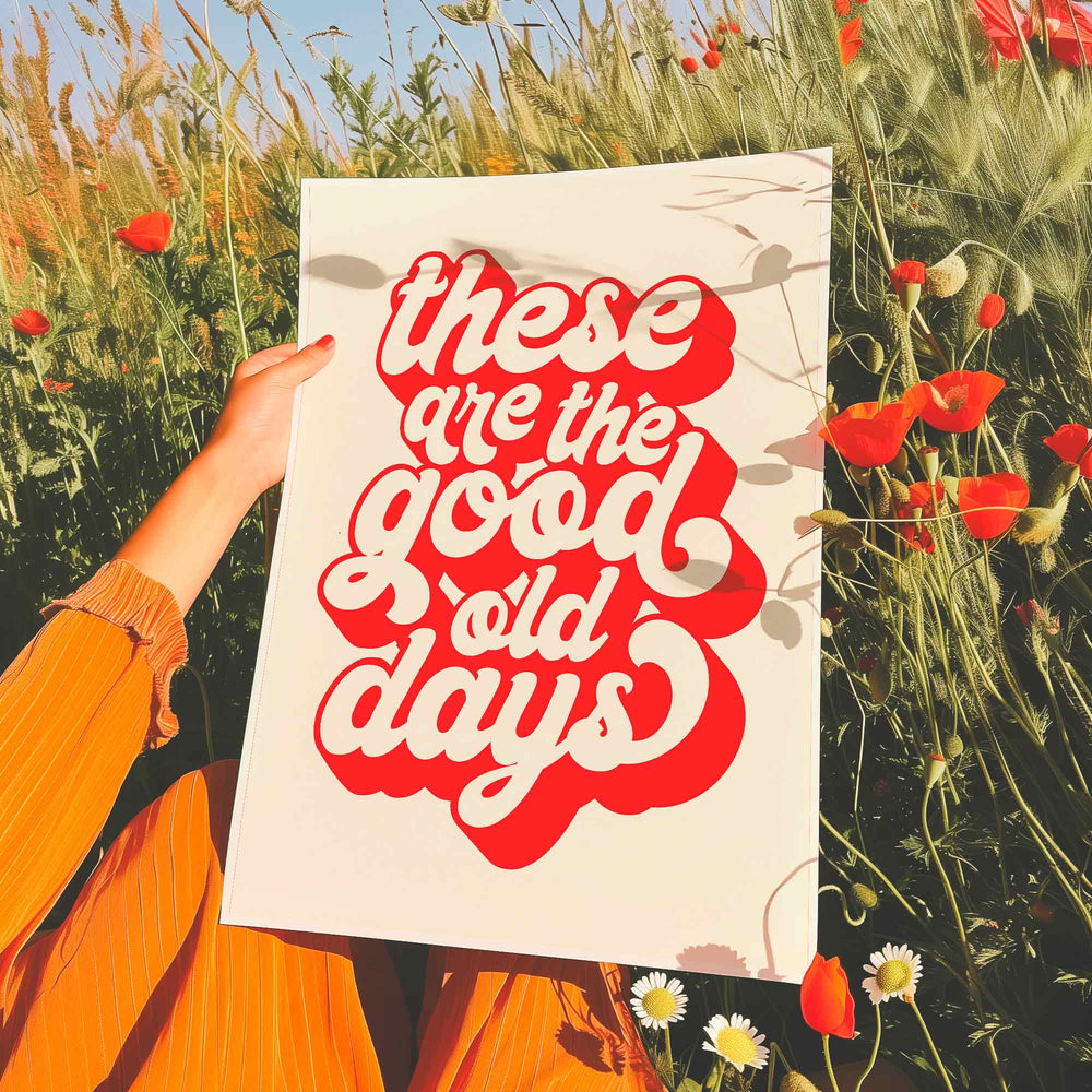 Retro-style motivational print held in a field of wildflowers, featuring bold red and white typography that reads “These are the good old days.”