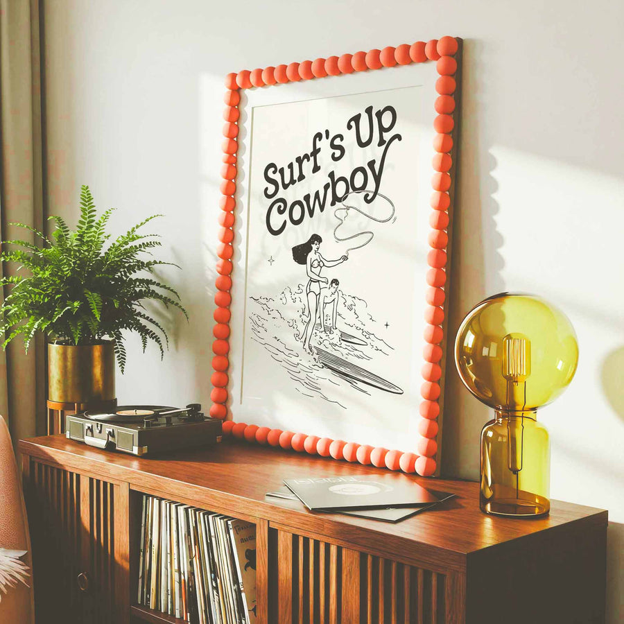 Surf’s Up Cowboy retro wall art print displayed in modern boho living room with orange beaded frame, mid-century wood console, record player, and yellow glass lamp.